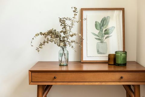 A mid-century wooden console styled with eucalyptus in a glass vase, a framed botanical print and a small candle.